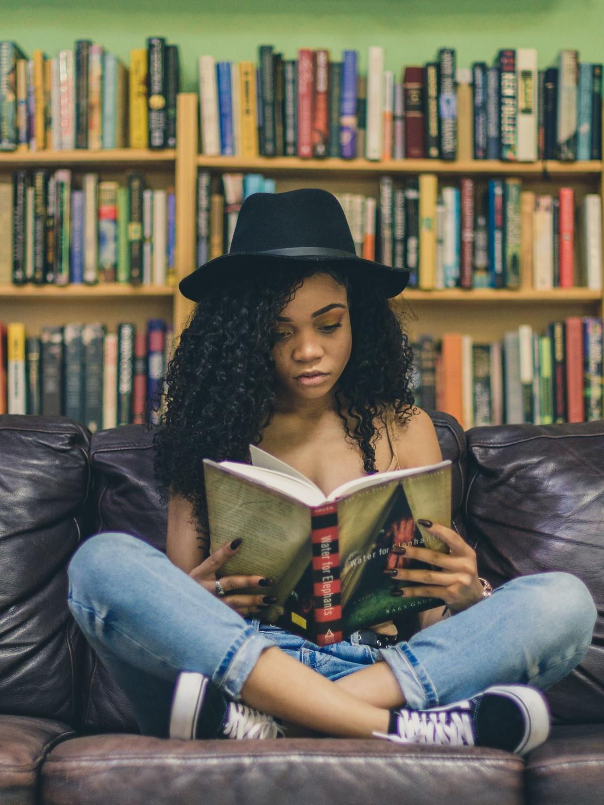 girl sitting on a sofa in front of a bookshelf, wearing a hate and reading a book